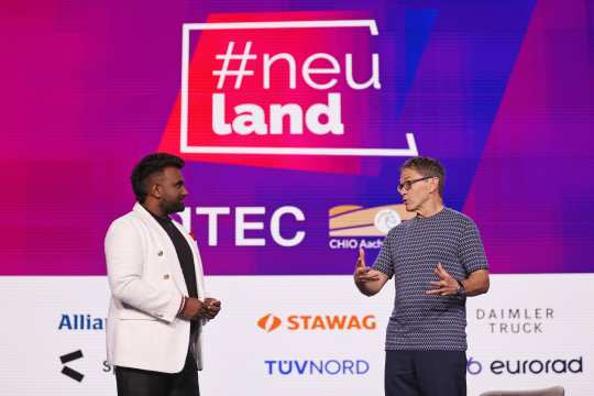 Bill Anderson auf der Bühne des Kongress #neuland vor einer großen Videowand mit Sponsoren stehend im Gespräch mit dem Interviewer.