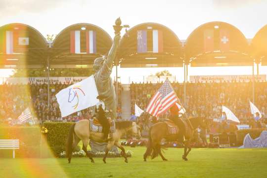 Two western riders on their horses riding through the sold-out main stadium in the evening sun during the opening ceremony, carrying a USA flag and a flag with the CHIO Aachen logo