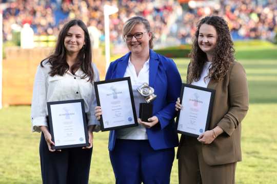 Three people are standing next to each other on a green lawn, each holding an award in their hands. Two of the people are wearing framed certificates with the inscription ‘Silver Camera’, while the person in the centre is also holding a silver trophy. They are formally dressed, and a grandstand and other people can be vaguely recognised in the background. The scene looks solemn and professional.