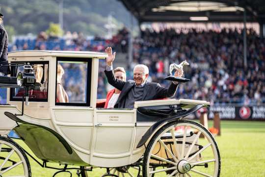 Eine elegante, cremefarbene Pferdekutsche fährt über eine grüne Rasenfläche, während ein Mann auf dem Rücksitz sitzend eine Trophäe in Form eines Pferdes hochhält. Die Kutsche ist mit einem Namensschild versehen, auf dem "Cremer Oberzier" steht. Im Hintergrund ist eine große Zuschauertribüne mit vielen Menschen zu sehen, die den CHIO Aachen verfolgen. Die Atmosphäre wirkt festlich und feierlich.