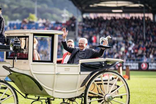 Eine elegante, cremefarbene Pferdekutsche fährt über eine grüne Rasenfläche, während ein Mann auf dem Rücksitz sitzend eine Trophäe in Form eines Pferdes hochhält. Die Kutsche ist mit einem Namensschild versehen, auf dem "Cremer Oberzier" steht. Im Hintergrund ist eine große Zuschauertribüne mit vielen Menschen zu sehen, die den CHIO Aachen verfolgen. Die Atmosphäre wirkt festlich und feierlich.