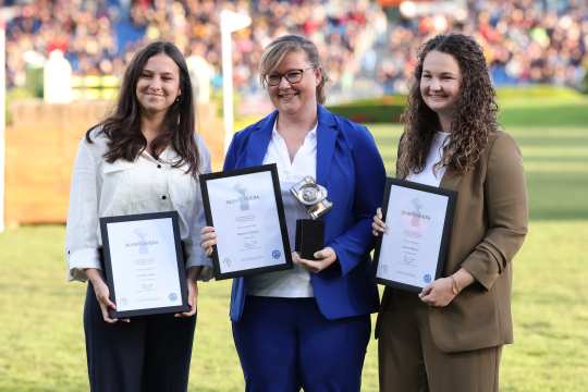 Three people are standing next to each other on a green lawn, each holding an award in their hands. Two of the people are wearing framed certificates with the inscription ‘Silver Camera’, while the person in the centre is also holding a silver trophy. They are formally dressed, and a grandstand and other people can be vaguely recognised in the background. The scene looks solemn and professional.