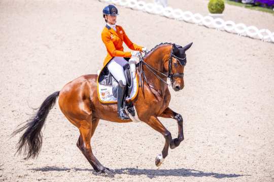 Dinja van Liere and Vita di Lusso performing a canter pirouette in the middle of the Dressage Stadium. Dinja wears an orange tailcoat and a blue helmet.